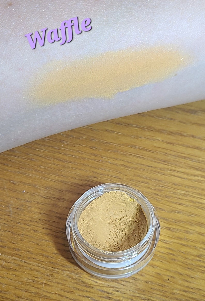 Puede incluir: Un peque&ntilde;o tarro de sombra de ojos de color amarillo dorado con la palabra "waffle" escrita en morado en el brazo de una persona.