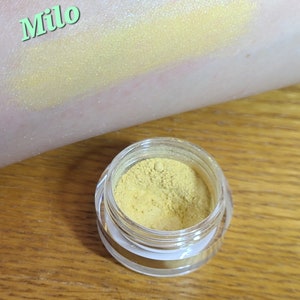 Puede incluir: Un peque&ntilde;o tarro de pigmento amarillo para sombra de ojos etiquetado como "Milo". El pigmento es un amarillo brillante y vibrante con un ligero brillo.