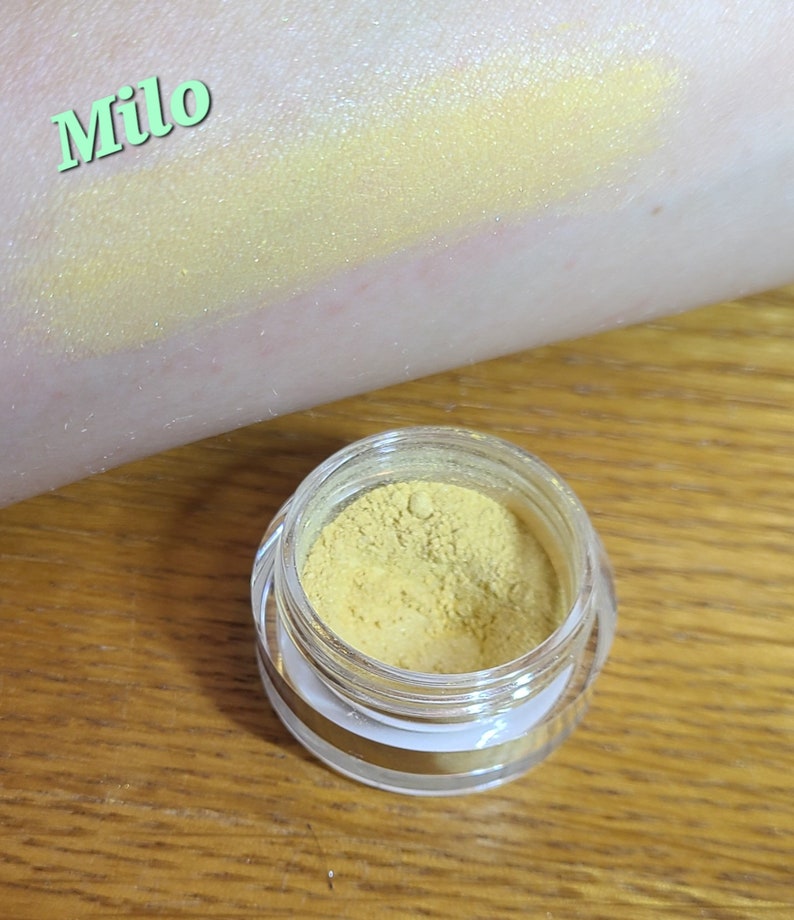 Puede incluir: Un peque&ntilde;o tarro de pigmento amarillo para sombra de ojos etiquetado como "Milo". El pigmento es un amarillo brillante y vibrante con un ligero brillo.