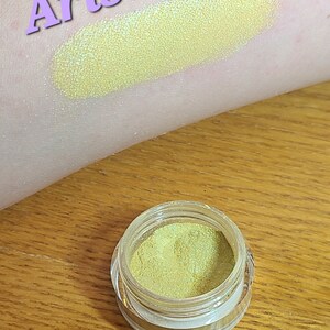 Puede incluir: Arista es el nombre de esta sombra de ojos amarilla con un acabado brillante. La sombra de ojos est&aacute; en un peque&ntilde;o frasco redondo con una tapa transparente.