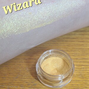 Puede incluir: Un peque&ntilde;o frasco de purpurina dorada etiquetado como "wizard". La purpurina se muestra en el brazo de una persona, creando un efecto brillante e iridiscente.