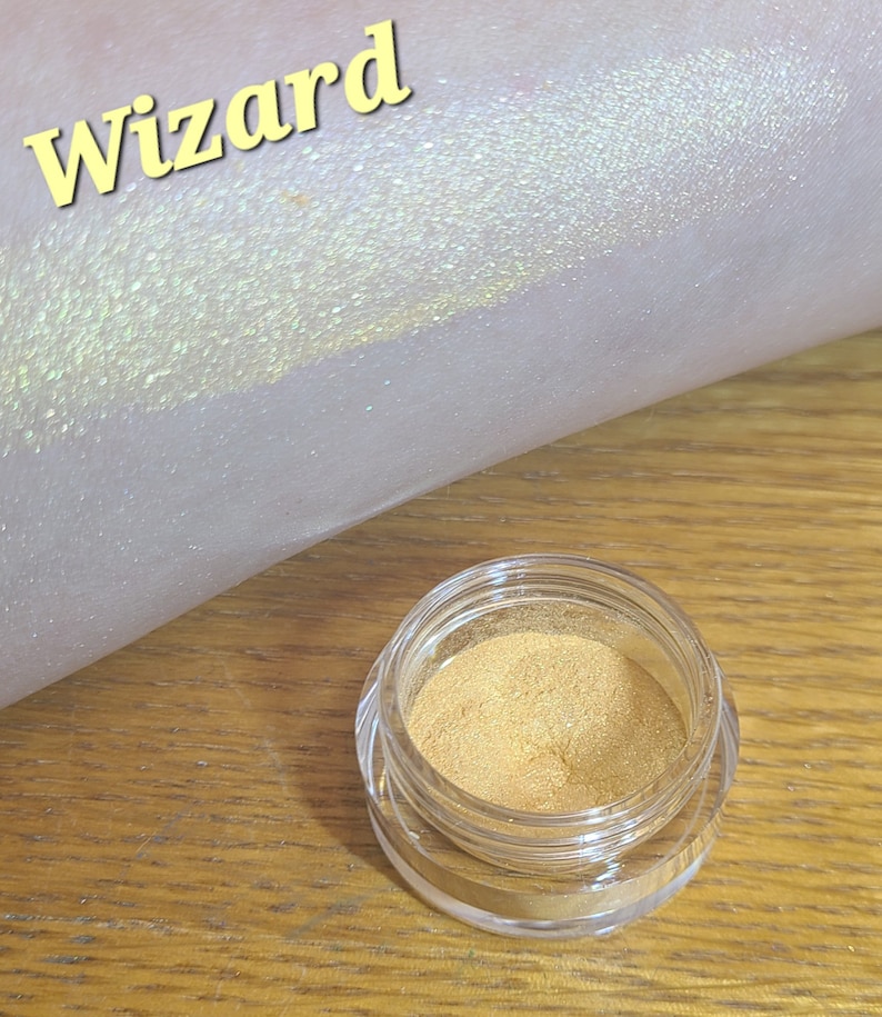 Puede incluir: Un peque&ntilde;o frasco de purpurina dorada etiquetado como "wizard". La purpurina se muestra en el brazo de una persona, creando un efecto brillante e iridiscente.