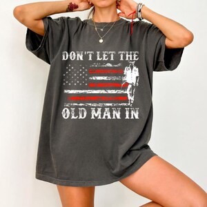 Könnte beinhalten: Dunkelgraues T-Shirt mit dem Text "DON'T LET THE OLD MAN IN" über einem amerikanischen Flaggen-Design und einer Silhouette. Das T-Shirt hat kurze Ärmel und eine lockere Passform. Maße in Zentimetern.