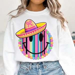 May include: White sweatshirt featuring a colorful smiley face design. The smiley face has a sombrero and a striped background with a variety of colors. The design is surrounded by small smiley faces in different colors.