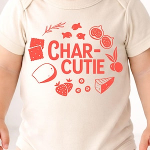 May include: A cream-colored baby onesie with a red graphic design. The design includes the text "CHAR-CUTIE" with illustrations of crackers, fish, figs, strawberries, cheese, and other food items.