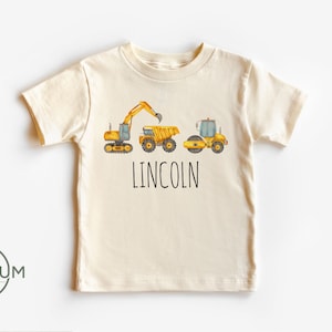 May include: Cream-colored t-shirt featuring a watercolor illustration of construction vehicles: an excavator, a dump truck, and a road roller. The name "LINCOLN" is printed in black block letters below the vehicles. The shirt is a children's apparel item.