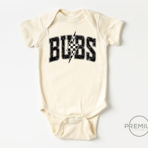 May include: Cream-colored baby onesie with the word "BUBS" in distressed black lettering. A black and white checkered lightning bolt graphic is centered within the text. The onesie has short sleeves and a snap closure.