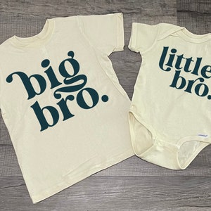 May include: Two cream colored t-shirts with the text "big bro." and "little bro." printed in dark teal on them. The shirts are on a wood background.