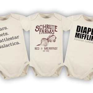 May include: Three white baby onesies with different designs. The first onesie has the text "Bears. Beets. Battlestar Galactica." The second onesie has the text "Schrute Farms Bed & Breakfast Est 1812" with a drawing of a beet. The third onesie has the text "Diaper Mifflin, Inc. Pooper Company."
