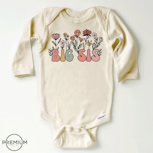 May include: A long-sleeved, cream-colored baby onesie with a graphic of the words "BIG SIS" in colorful, retro-style lettering surrounded by pink, green, and orange flowers.