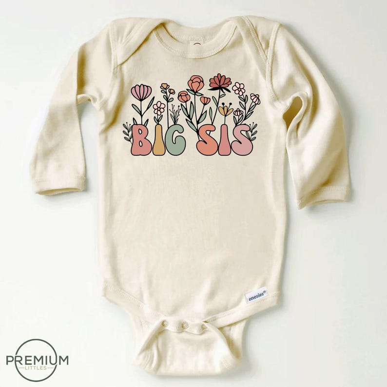 May include: A long-sleeved, cream-colored baby onesie with a graphic of the words "BIG SIS" in colorful, retro-style lettering surrounded by pink, green, and orange flowers.