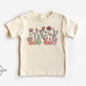 May include: A cream colored t-shirt with short sleeves featuring the text "BIG SIS" in colorful letters surrounded by pink, orange, and yellow flowers.