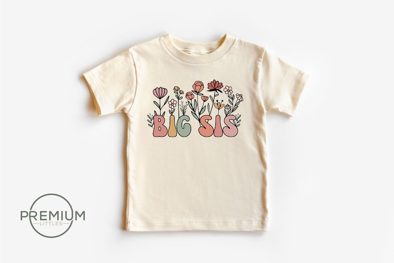 May include: A cream colored t-shirt with short sleeves featuring the text "BIG SIS" in colorful letters surrounded by pink, orange, and yellow flowers.
