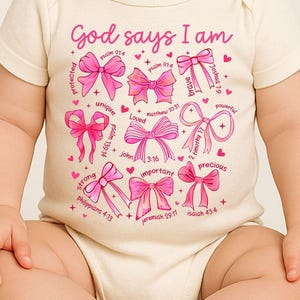 May include: Cream-colored baby bodysuit with the text "God says I am" and pink bow illustrations. The design includes words like "protected," "unique," "brave," and "precious," with Bible verses. A cute and inspirational baby garment.