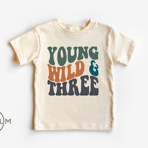 May include: A cream colored t-shirt with the text "YOUNG WILD & THREE" in retro-style font. The text is in shades of green, orange, and blue.