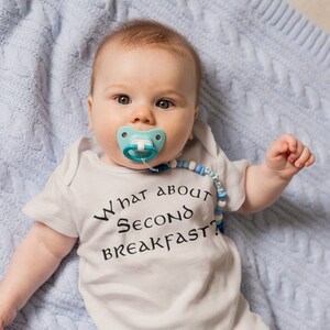 May include: A baby wearing a white onesie with the text "What about second breakfast?" in black. The baby is lying on a blue knitted blanket and has a blue and white dummy in their mouth.