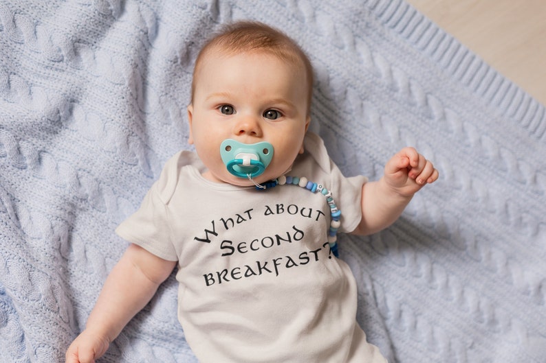 May include: A baby wearing a white onesie with the text "What about second breakfast?" in black. The baby is lying on a blue knitted blanket and has a blue and white dummy in their mouth.
