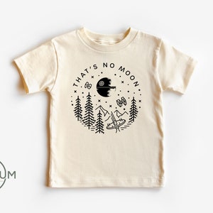 May include: A beige short-sleeved t-shirt with a black graphic design of a Death Star, a TIE fighter, and trees. The text "That's no moon" is written around the graphic.