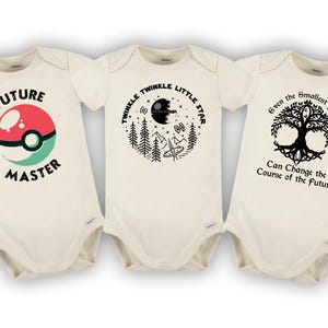 May include: Three white baby onesies with black text and graphics. The first onesie says "Future Master" with a red and green Pokeball graphic. The second onesie says "Twinkle Twinkle Little Star" with a black and white graphic of a star and a forest. The third onesie says "Even the smallest person can change the course of the future" with a black and white tree graphic.