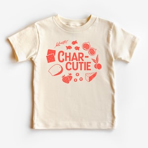 May include: Cream-colored toddler t-shirt with a red graphic. The graphic features the word "CHAR-CUTIE" surrounded by illustrations of food items like figs, strawberries, cheese, and crackers. The shirt has short sleeves and a crew neck.