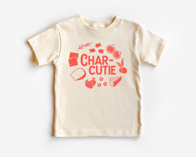 May include: Cream-colored toddler t-shirt with a red graphic. The graphic features the word "CHAR-CUTIE" surrounded by illustrations of food items like figs, strawberries, cheese, and crackers. The shirt has short sleeves and a crew neck.