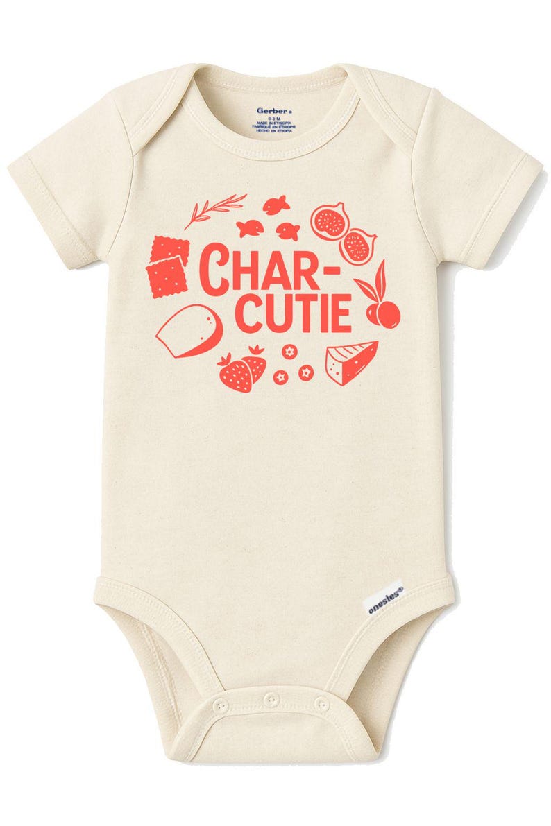 May include: Cream-colored baby onesie with short sleeves. The front features the red text "CHAR-CUTIE" with illustrations of crackers, fish, figs, cheese, strawberries, and other food items. The onesie has a snap closure.