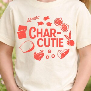 May include: Cream-colored t-shirt with a red graphic design. The design includes the text "CHAR-CUTIE" surrounded by illustrations of crackers, fish, figs, strawberries, cheese, and other food items.