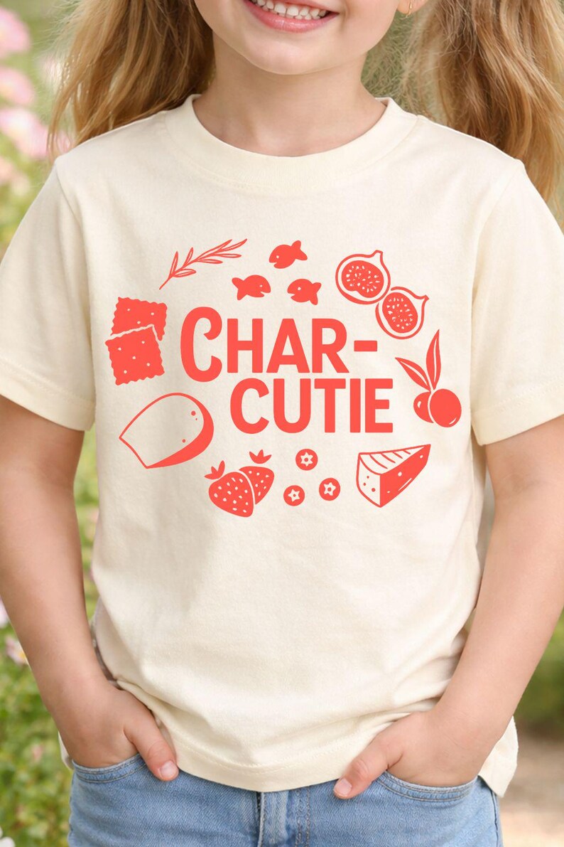 May include: Cream-colored t-shirt with a red graphic design. The design includes the text "CHAR-CUTIE" surrounded by illustrations of crackers, fish, figs, strawberries, cheese, and other food items.