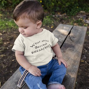 May include: A toddler wearing a white t-shirt with the text "What about second breakfast?" in black. The toddler is sitting on a wooden bench outdoors.