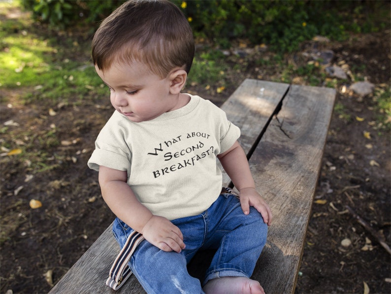 May include: A toddler wearing a white t-shirt with the text "What about second breakfast?" in black. The toddler is sitting on a wooden bench outdoors.