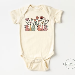 May include: A cream-colored baby onesie with a floral design and the text "BIG SIS" in colorful, stylized letters.