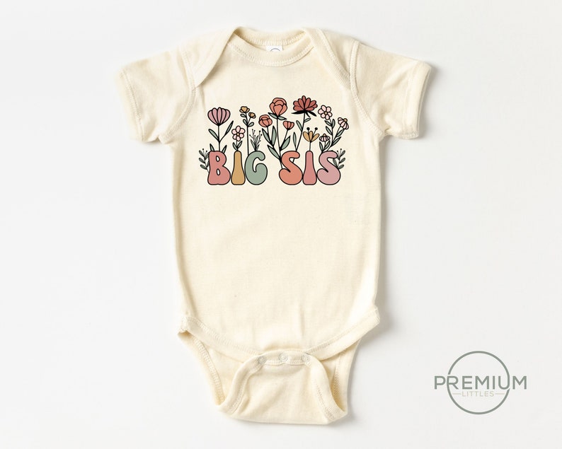 May include: A cream-colored baby onesie with a floral design and the text "BIG SIS" in colorful, stylized letters.