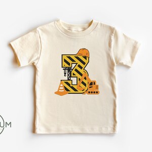 May include: A cream colored t-shirt with a yellow and black construction themed number 3. The number is decorated with a construction worker's hat, a construction cone, and a small excavator.