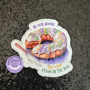 May include: A white sticker with a purple border features a cartoon illustration of a donut with purple, green, and gold sprinkles. The donut is dripping with white icing. The text "In our house the knife stays in the box" is written in green and purple letters.