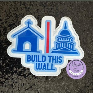May include: A blue and white sticker with a church, a red stripe, and the Capitol building. The sticker says "Build This Wall" and has a small logo that says "Hassle Free Creations".