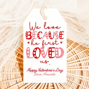 May include: A white gift tag with a string, featuring red and pink text that reads "We love BECAUSE he first LOVED us." Below, it says "Happy Valentine's Day from Amanda."