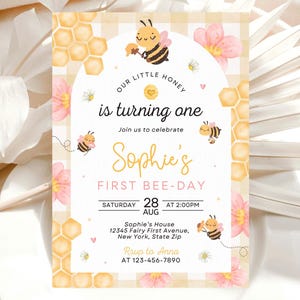May include: A whimsical invitation with a bee theme. The design features cartoon bees, honeycomb patterns, and pink flowers. The text reads "Our Little Honey is turning one" and announces "Sophie's First Bee-Day" with date and time details.
