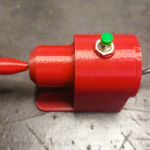 May include: A red 3D-printed cylindrical device featuring a small, protruding red nozzle at the front and a bright green circular push-button switch mounted on the top. A thin, dark wire extends from the back of the main red housing unit.