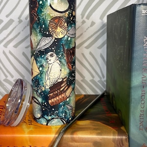 Puede incluir: Un vaso de acero inoxidable con un diseño temático de Harry Potter. El vaso presenta un fondo azul y verde con imágenes de gafas, un búho nival, una snitch dorada, un sombrero y libros. El vaso está colocado encima de un libro de Harry Potter.