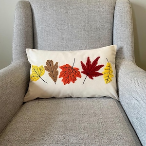 May include: A cream rectangular cushion featuring embroidered autumn leaves in yellow, brown, orange, and red. The cushion is placed on a grey armchair, creating a cosy, seasonal aesthetic. The leaves have black stems.