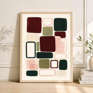 May include: Abstract art print in a light wood frame. The artwork features a collection of overlapping, rounded rectangles in shades of burgundy, olive green, dark green, pink, and cream against a cream background. The art is displayed on a wooden surface.