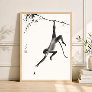 May include: A framed black and white ink wash painting of a monkey hanging from a tree branch. The monkey reaches for a spider. The artwork has Chinese characters and a red stamp in the corner. The frame is a light wood color.