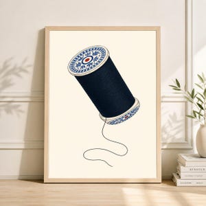 Op de afbeelding: Een ingelijste print met een gestileerde illustratie van een spoel donkerblauw garen. De spoel heeft decoratieve witte en blauwe details aan de boven- en onderkant. Een enkele draad loopt uit de spoel tegen een crèmekleurige achtergrond.