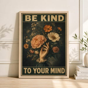 May include: A framed print with the text "BE KIND TO YOUR MIND". The artwork features a hand holding a bouquet of orange, peach, and white flowers against a dark green background. The frame is a light wood color.