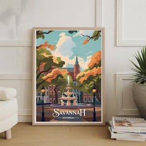 Op de afbeelding: Een ingelijste kunstprint met een gestileerde illustratie van een fontein in Savannah, Georgia. Het kunstwerk toont een fontein met opspuitend water, omringd door bomen met oranje en groene bladeren, en de tekst "Savannah Georgia".