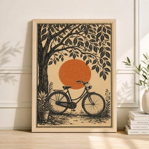 May include: A framed art print featuring a black and white illustration of a bicycle parked under a tree with a large orange sun in the background. The artwork has a vintage aesthetic, with a potted plant on the left.