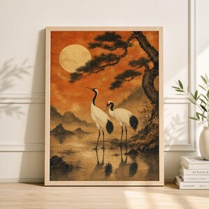 Könnte beinhalten: Gerahmter Kunstdruck mit zwei japanischen Kranichen in einer ruhigen Landschaft. Das Kunstwerk zeigt einen Vollmond, einen großen Baum und Berge, alles in warmen Orange- und Brauntönen. Die Kraniche haben weiße Körper, schwarze Flügelspitzen und rote Kopfmarkierungen.
