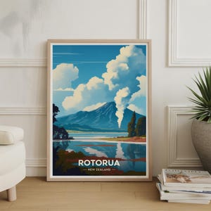 以下が含まれることがあります： ニュージーランド、ロトルアの風景を描いた額入りのアートプリント。山、湖、雲が青い空の下に描かれています。プリントには、白い文字で「ROTORUA - NEW ZEALAND」と書かれています。