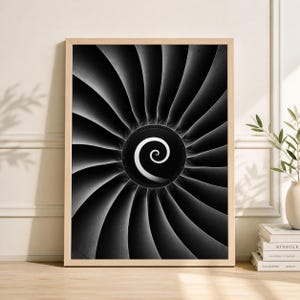 Puede incluir: Fotografía en blanco y negro enmarcada de una turbina de motor a reacción. La imagen presenta un diseño en espiral en el centro, rodeado de aspas curvas. La obra de arte se muestra en un marco de madera de color claro, creando una estética moderna.