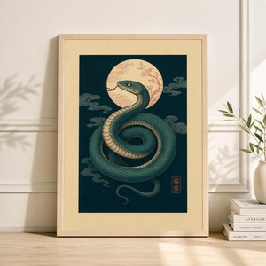 May include: Framed art print featuring a teal snake coiled in front of a full moon with cherry blossoms. The artwork has a vintage aesthetic with a dark blue background and stylized clouds. A small red and gold square with Japanese text is in the lower right corner.